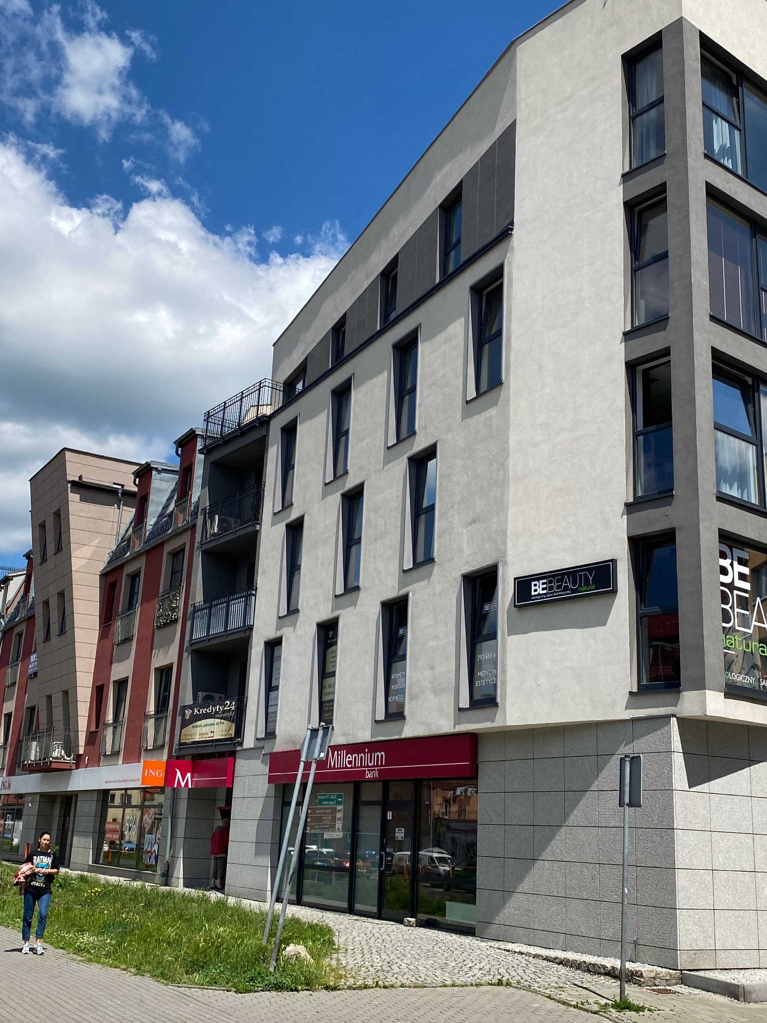 Nowoczesny budynek z lokalami usługowymi na parterze, w tym bank Millennium i salon kosmetyczny BE Beauty, widoczne reklamy i szyldy, chodnikiem przechodzi kobieta.