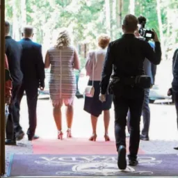 Kamerzysta z profesjonalnym sprzętem filmuje wchodzących gości na czerwonym dywanie, widok zza otwartych drzwi wejściowych z logo lokalu.