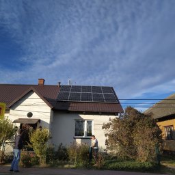 PV-Power - Dom jednorodzinny z brązowym dachem i panelami fotowoltaicznymi na dachu, widoczni dwaj mężczyźni na zewnątrz budynku, błękitne niebo z chmurami w tle, widoczny znak drogowy z sylwetką dziecka.