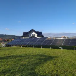 Instalacja paneli słonecznych na trawniku przed domem jednorodzinnym z czarnym dachem w słoneczny dzień.