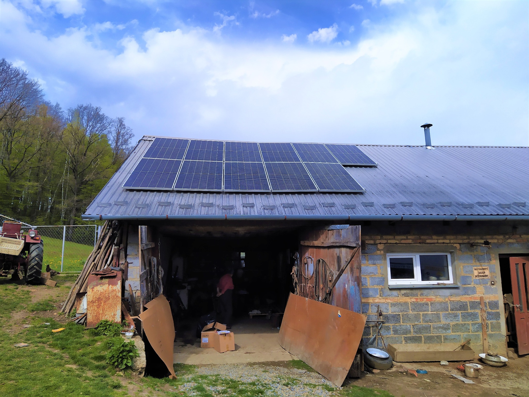 Instalacja paneli fotowoltaicznych na dachu budynku gospodarczego z widocznym traktorem w tle, częściowo otwarte wrota garażowe i niebieskie niebo z chmurami.