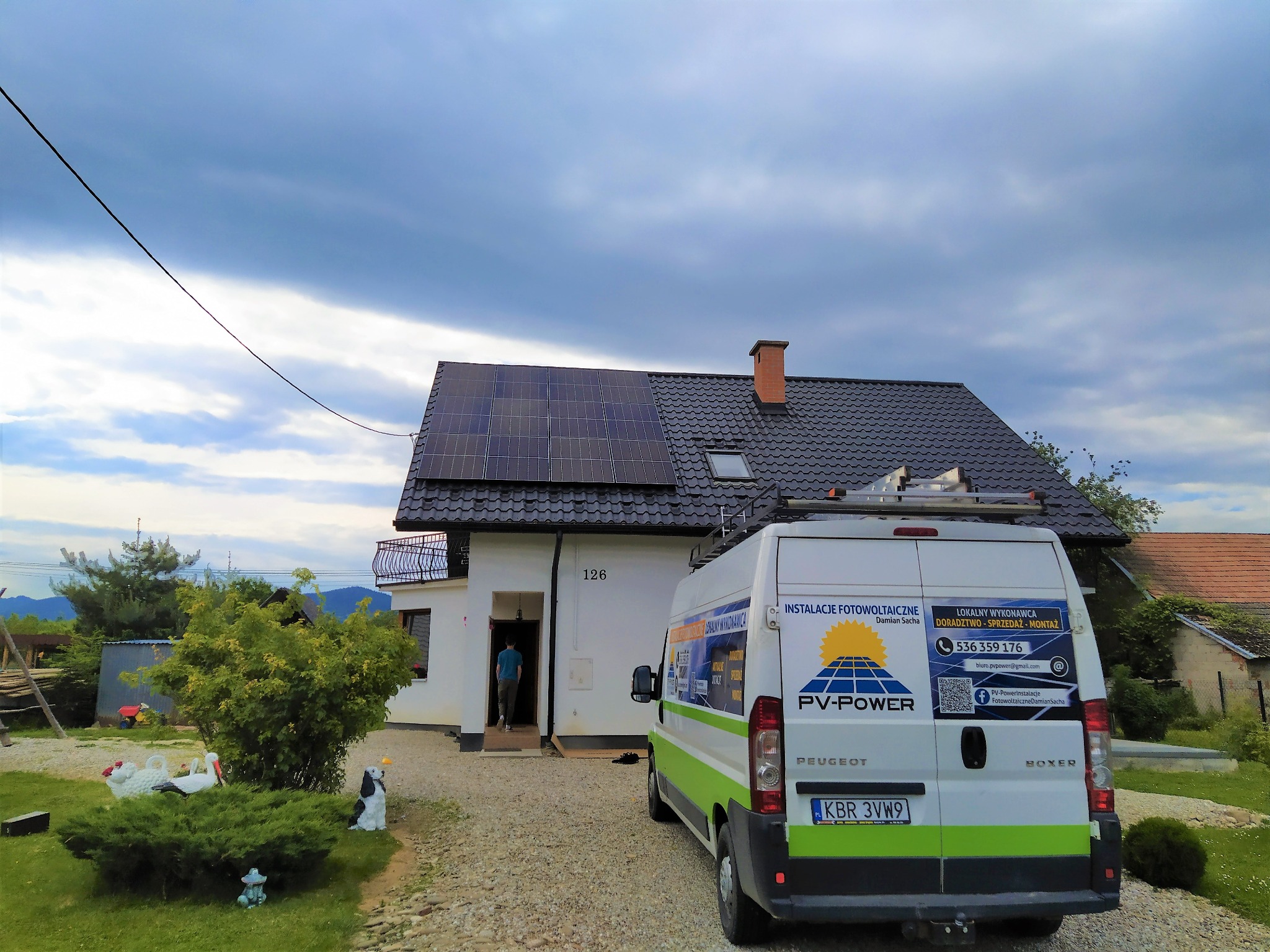 Dom jednorodzinny z panelami słonecznymi na dachu, zaparkowany biały bus z reklamą instalacji fotowoltaicznych, widoczny numer domu 126 i osoba wchodząca do środka.
