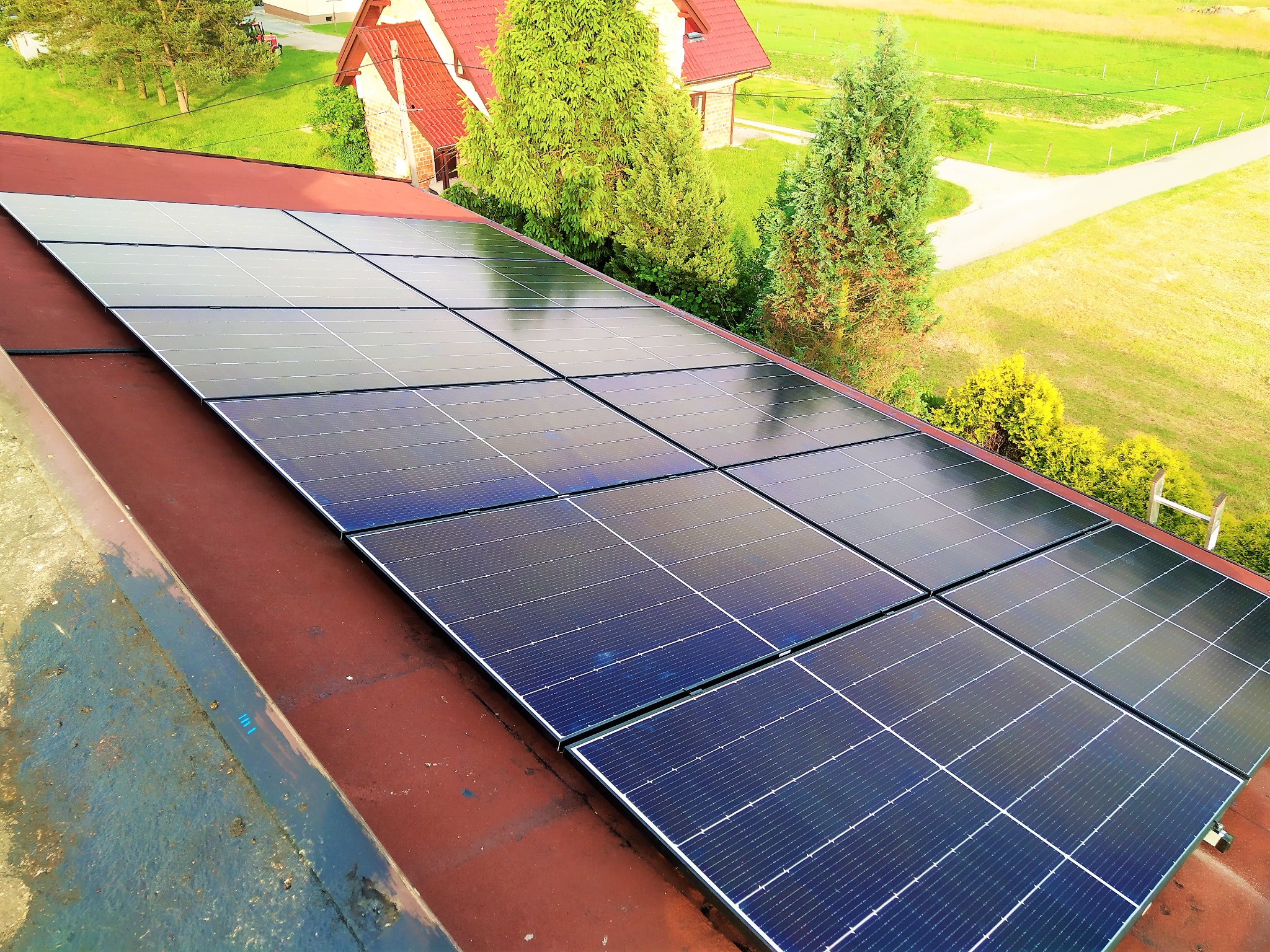 Panele słoneczne zamontowane na czerwonym dachu domu jednorodzinnego, widok z góry, w tle zieleń drzew i trawnik.