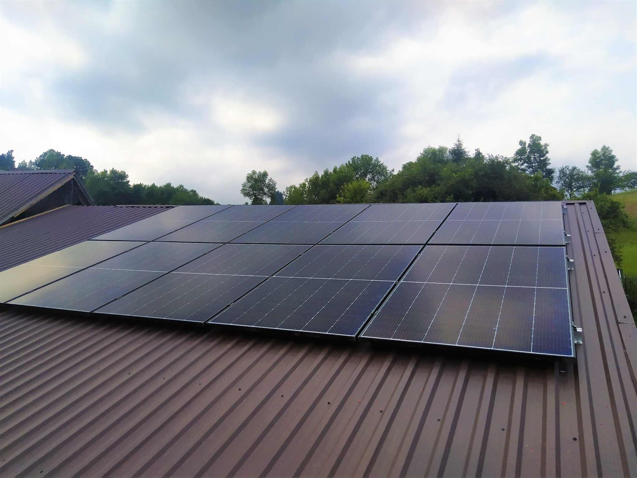 Panele słoneczne zamontowane na brązowym, metalowym dachu domu jednorodzinnego, widok na pochmurne niebo i zieleń drzew w tle.