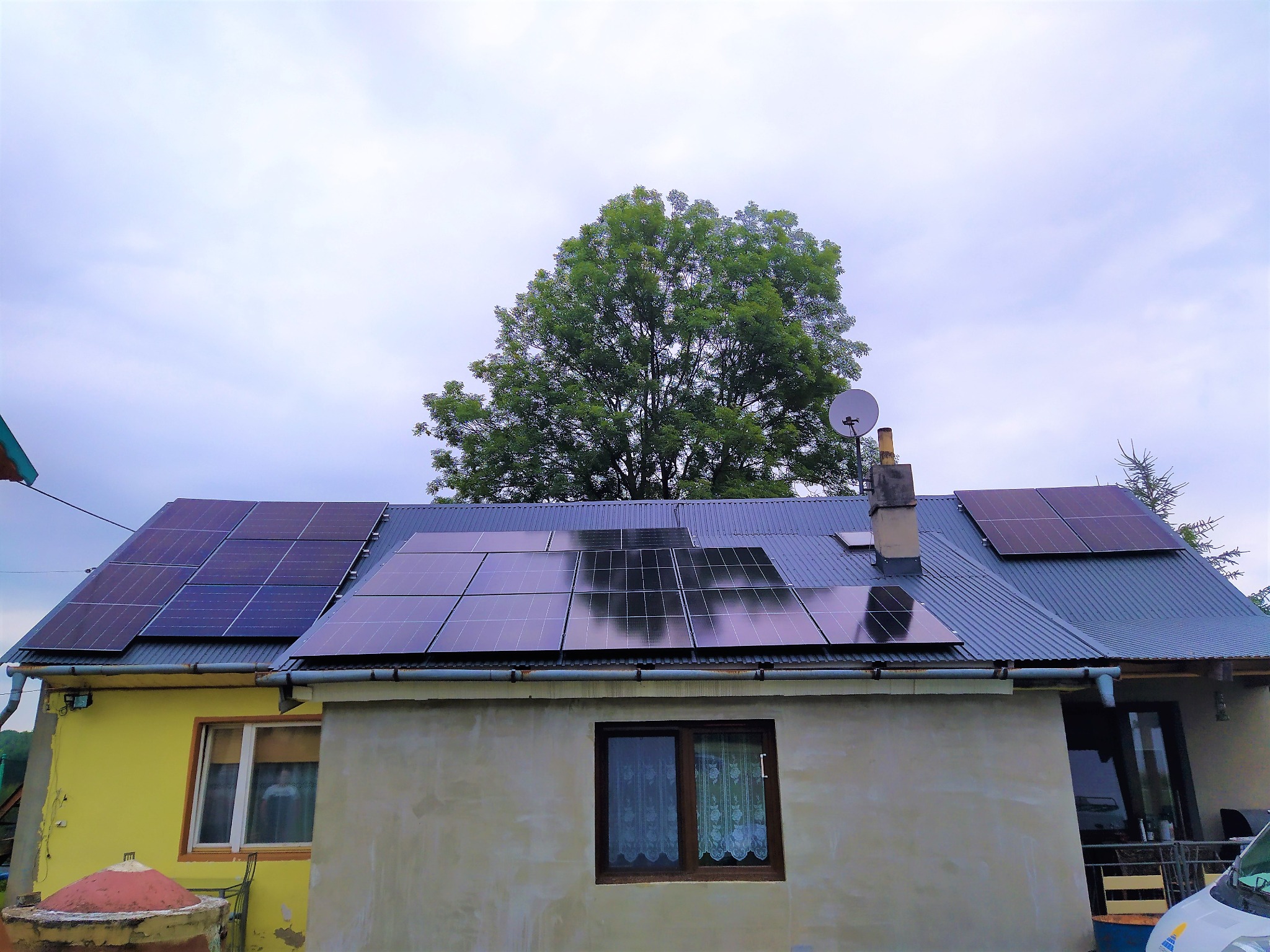 Dom jednorodzinny z panelami słonecznymi na dachu z blachy falistej, widok z dołu na tle zielonego drzewa i pochmurnego nieba.