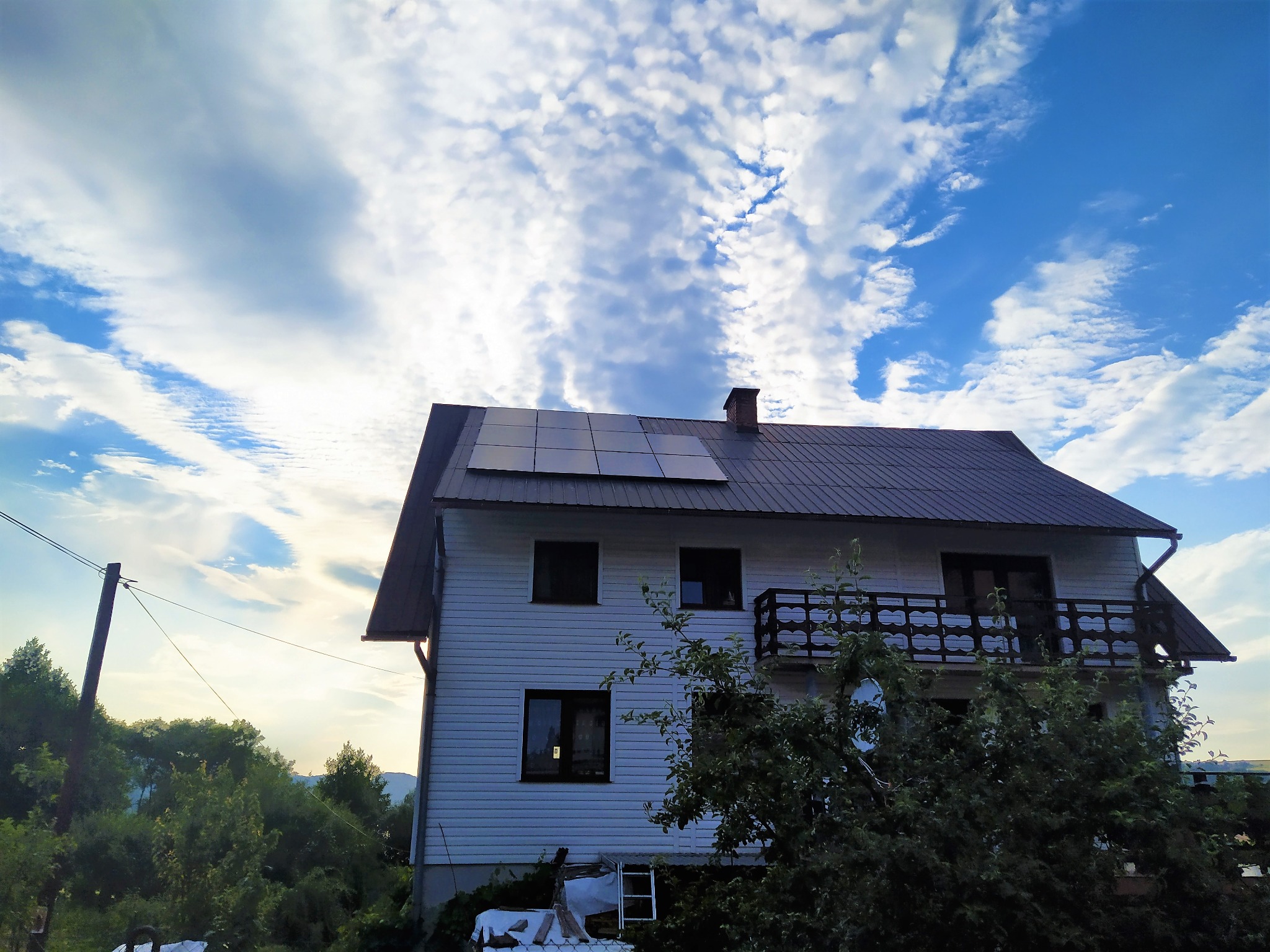 Biały dom dwupiętrowy z ciemną dachówką i panelami słonecznymi, widok z dołu na tle błękitnego nieba z chmurami, otoczony zielenią.