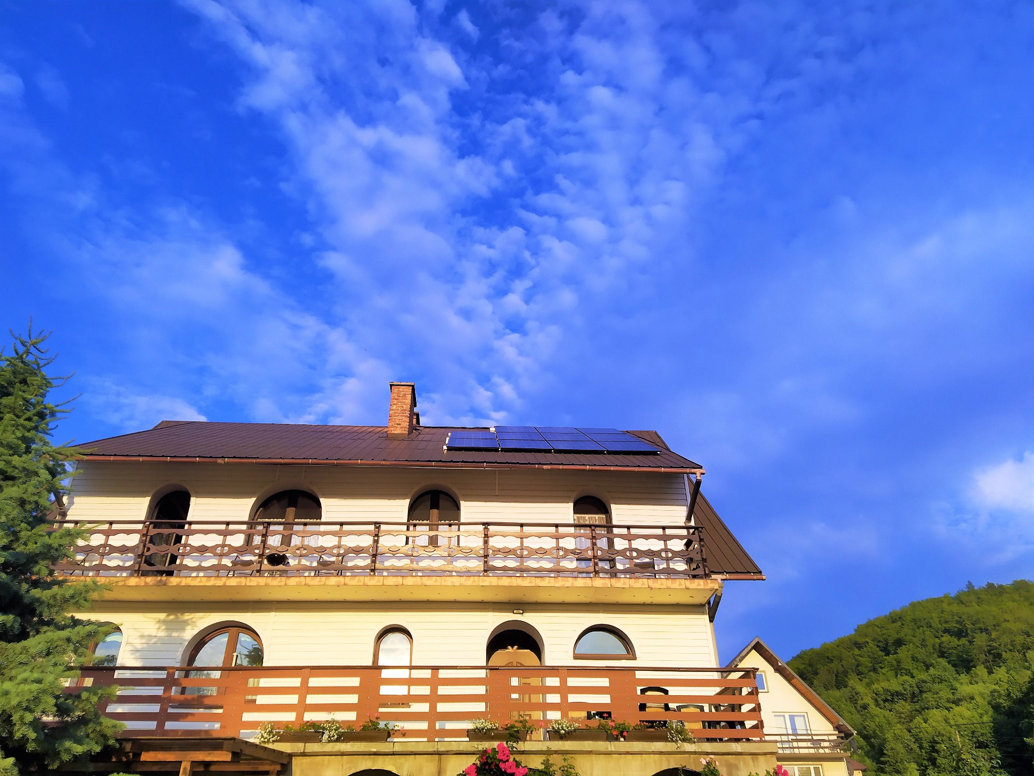 Dwupiętrowy dom z brązowym dachem, balkonami z drewnianą balustradą i panelami słonecznymi na dachu, na tle błękitnego nieba z chmurami i zielonego wzgórza.