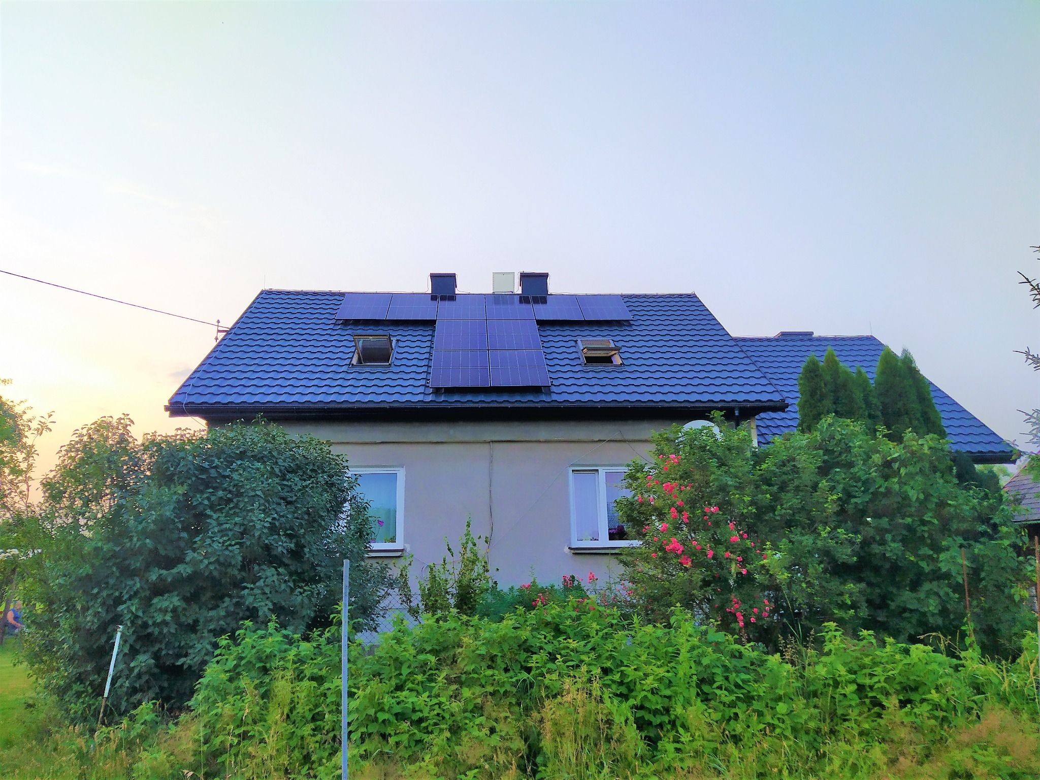 Dom jednorodzinny z niebieską dachówką, na której zamontowane są panele słoneczne; widoczne okna dachowe i kominy, a przed domem bujna roślinność.