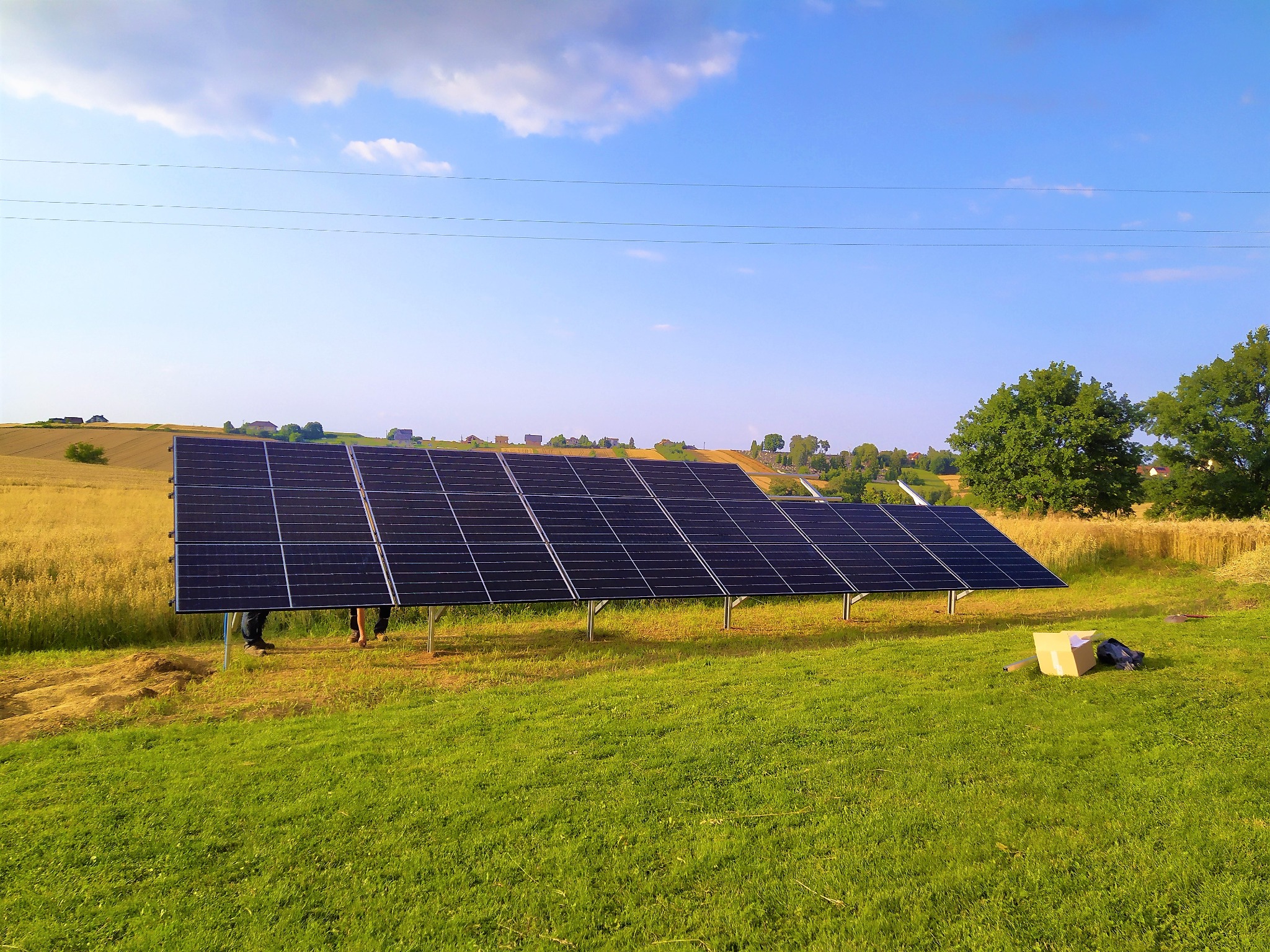 Panele fotowoltaiczne w trakcie montażu na łące, widoczne nogi instalatorów pod konstrukcją, w tle pole zboża i błękitne niebo.