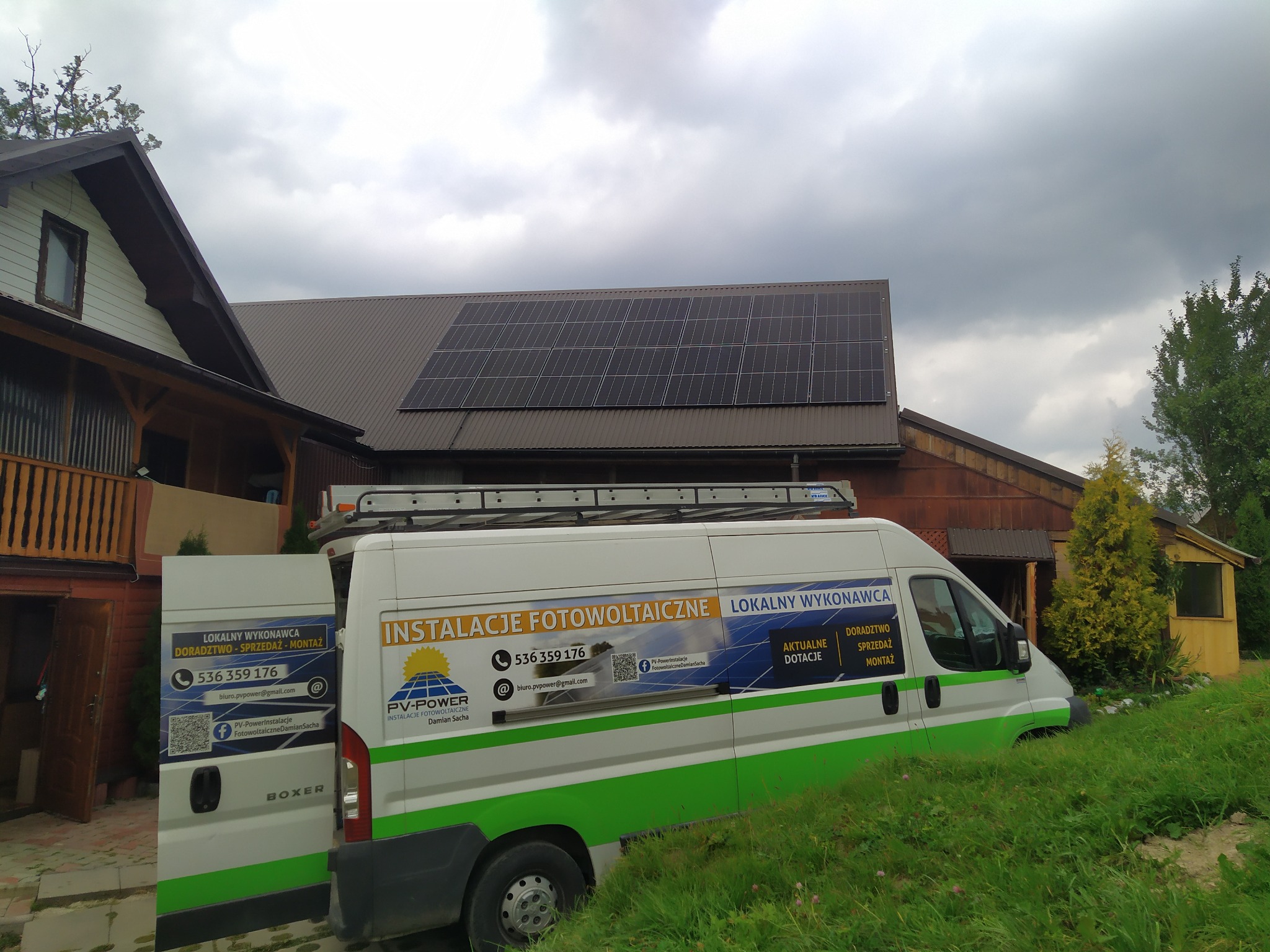 Instalacja paneli słonecznych na dachu brązowego budynku mieszkalnego, widok z perspektywy podjazdu, z zaparkowanym białym busem z zielonym pasem i reklamą firmy instalującej systemy fotowoltaiczne.