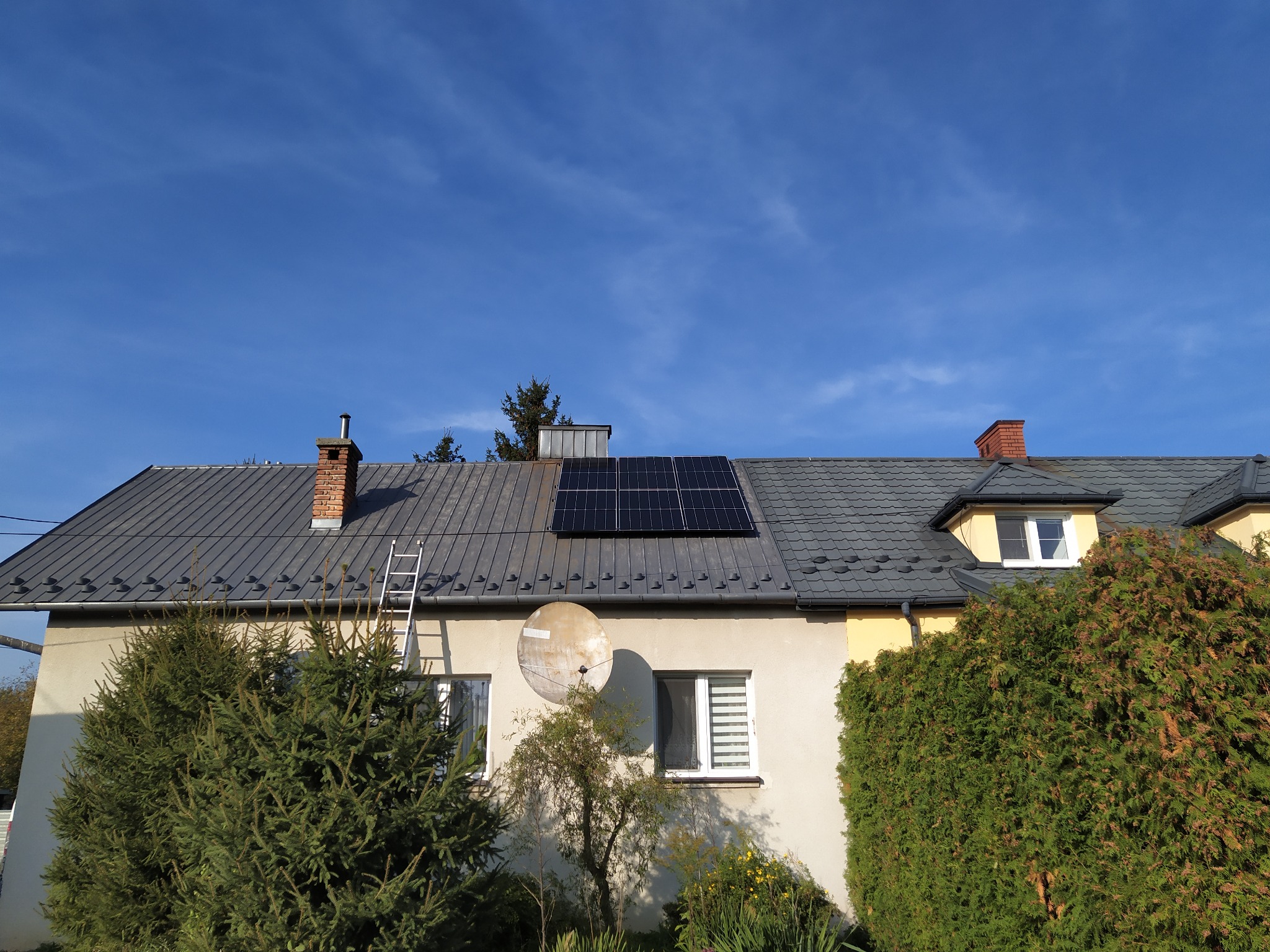 Panele słoneczne na dachu domu jednorodzinnego z szarą dachówką, widoczne z poziomu ogrodu, z anteną satelitarną na elewacji i drabiną opartą o ścianę.