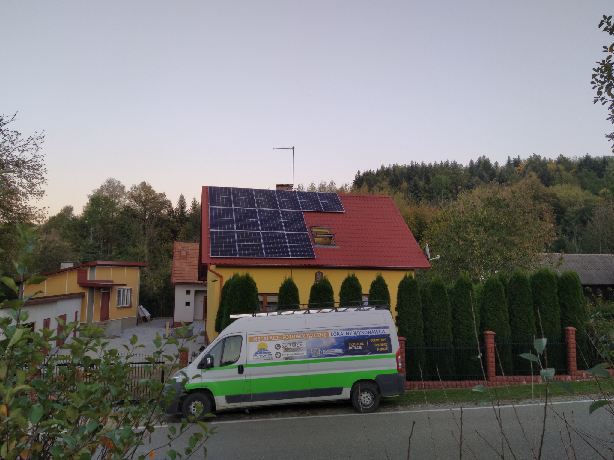 Dom jednorodzinny z czerwoną dachówką i panelami słonecznymi na dachu, na tle drzew, z zaparkowanym samochodem dostawczym firmy instalującej fotowoltaikę.
