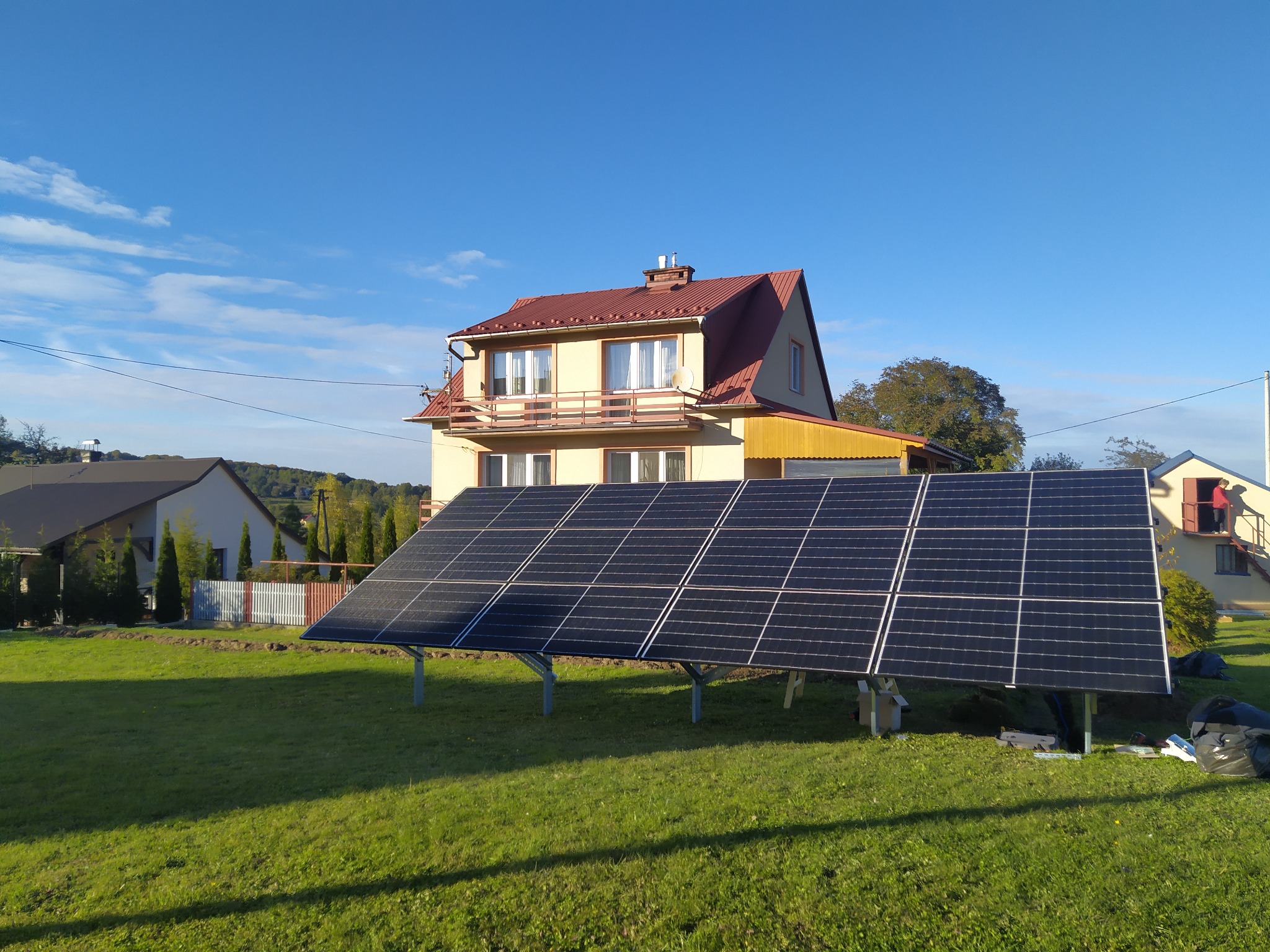 Panele słoneczne na trawniku przed domem jednorodzinnym, widoczne narzędzia i materiały montażowe, błękitne niebo w tle.