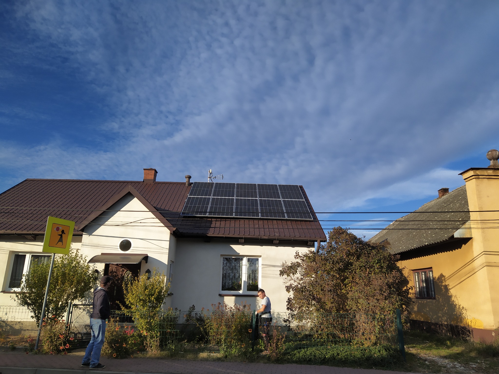 Dom jednorodzinny z brązowym dachem i panelami fotowoltaicznymi na dachu, widoczni dwaj mężczyźni na zewnątrz budynku, błękitne niebo z chmurami w tle, widoczny znak drogowy z sylwetką dziecka.