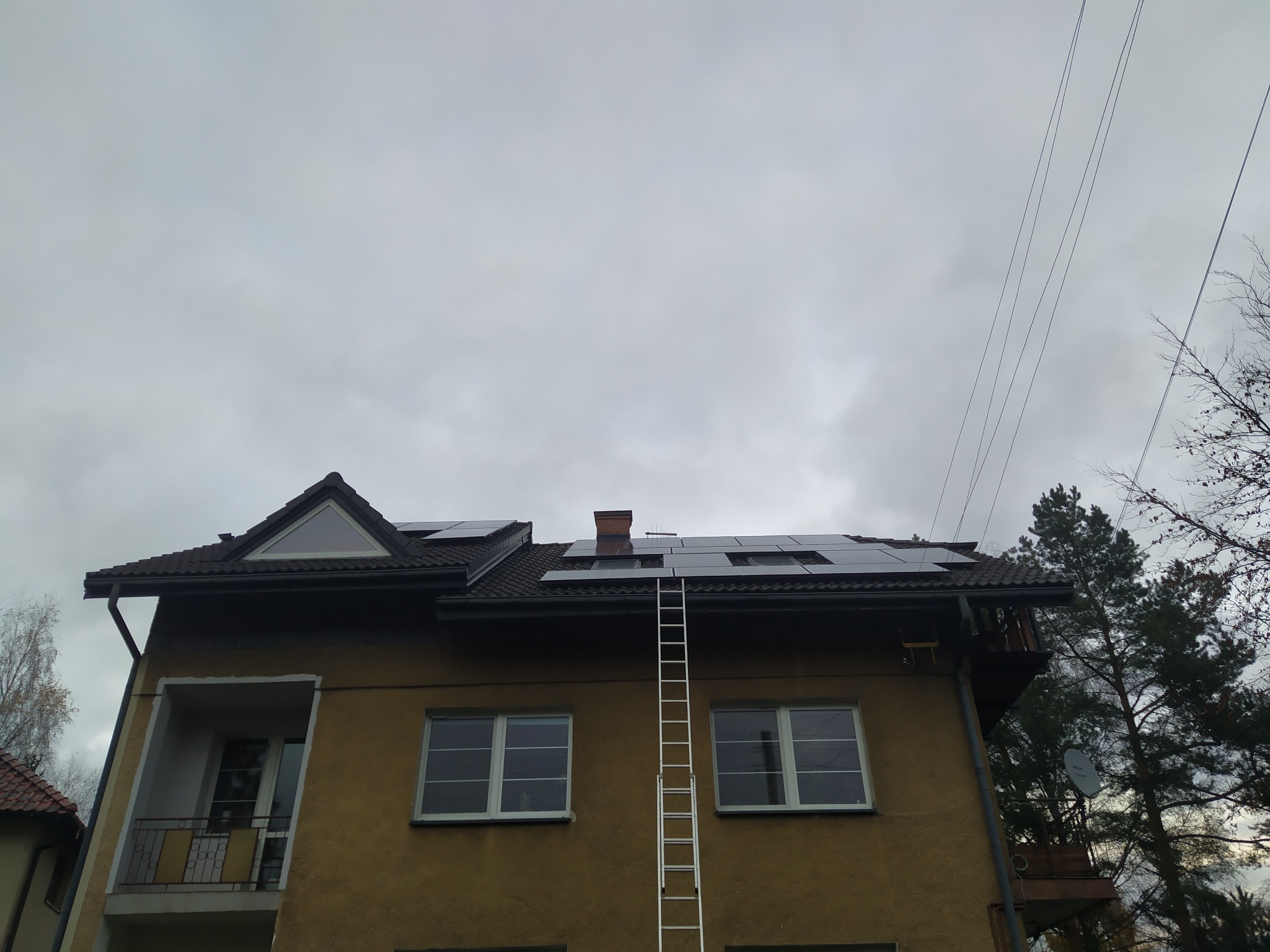 Montaż paneli fotowoltaicznych na dachu domu jednorodzinnego podczas pochmurnego dnia, drabina oparta o ścianę budynku.