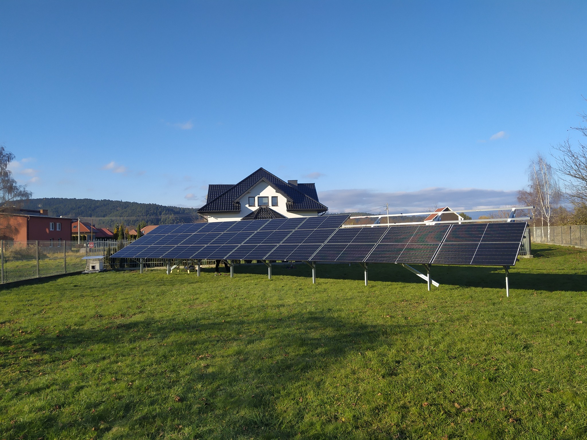 Instalacja paneli słonecznych na trawniku przed domem jednorodzinnym z czarnym dachem w słoneczny dzień.