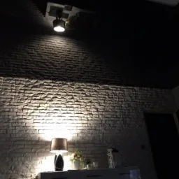 Ściana z dekoracyjnej cegły oświetlona lampą stojącą i punktowym światłem sufitowym, nad komodą z dekoracjami, w pomieszczeniu z czarnym sufitem.