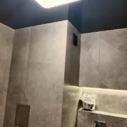 Wykończona łazienka z szarymi płytkami imitującymi marmur, podświetleniem LED pod sufitem, czarnym sufitem i dekoracyjną metalową miniaturą wanny.