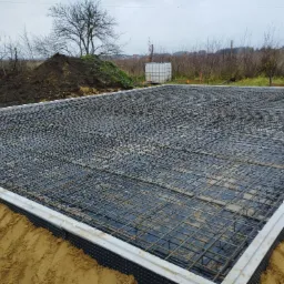 Zbrojenie fundamentu w siatce z prętów stalowych na czarnej folii hydroizolacyjnej, z widocznymi szalunkami i rurami wentylacyjnymi, na tle wykopu i krajobrazu z drzewami bezlistnymi.