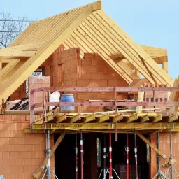 Surowy stan budynku z cegły ceramicznej z drewnianą konstrukcją dachu w trakcie budowy, widoczne rusztowania i podpory.
