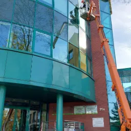 Pracownik w pomarańczowym podnośniku koszowym myje okna nowoczesnego biurowca z zieloną elewacją i ceglanym narożnikiem, widoczne odbicia drzew w szybach.