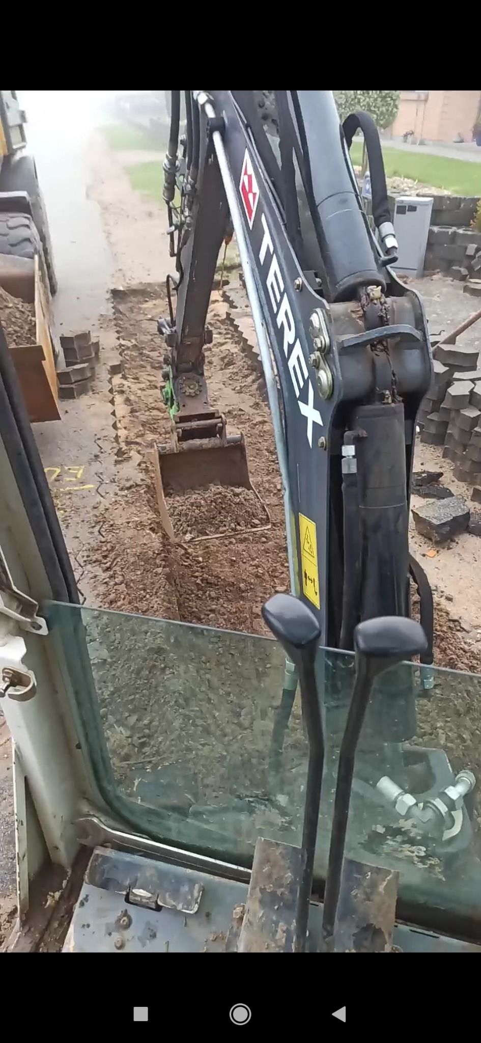 Widok z kabiny koparki Terex na wykop pod instalacje, w tle kostka brukowa i droga.