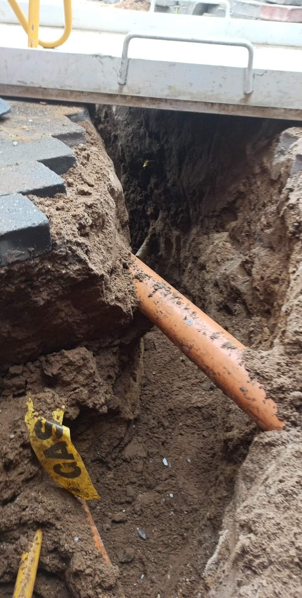 Pomarańczowa rura gazowa ułożona w wykopie ziemnym, widoczny żółty znacznik gazu i krawędź chodnika z kostki brukowej.