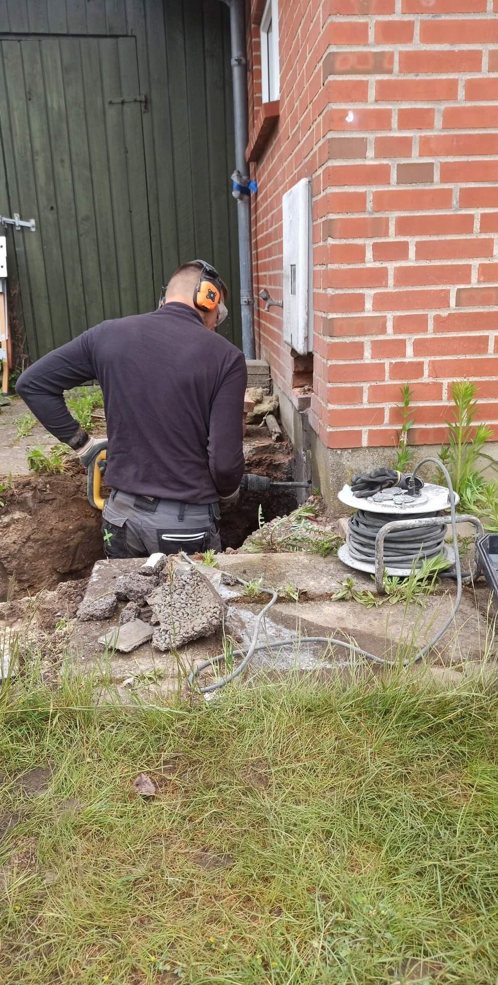 Pracownik w słuchawkach wierci wiertarką udarową dziurę w ziemi przy fundamencie ceglanego budynku, obok leży zwinięty przedłużacz i gruz.