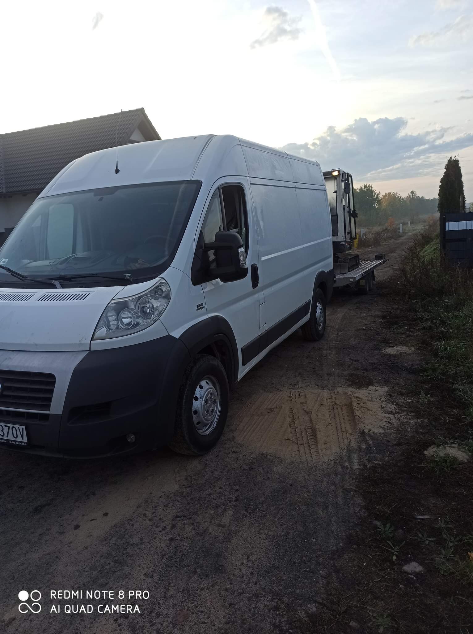 Biały bus z przyczepą przewożącą małą koparkę na błotnistej drodze dojazdowej, widoczny fragment domu w tle, pochmurne niebo.