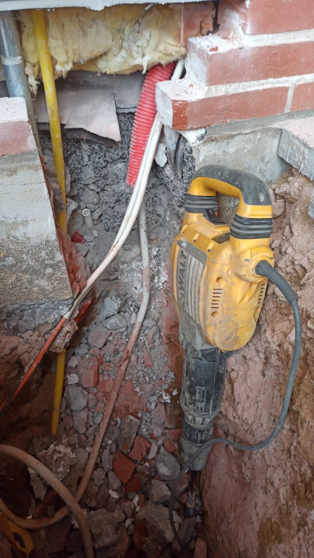 Żółty młot udarowy DeWalt w wykopie z gruzem ceglanym i kablami, widoczne ślady użycia, fragment muru z cegły w tle.