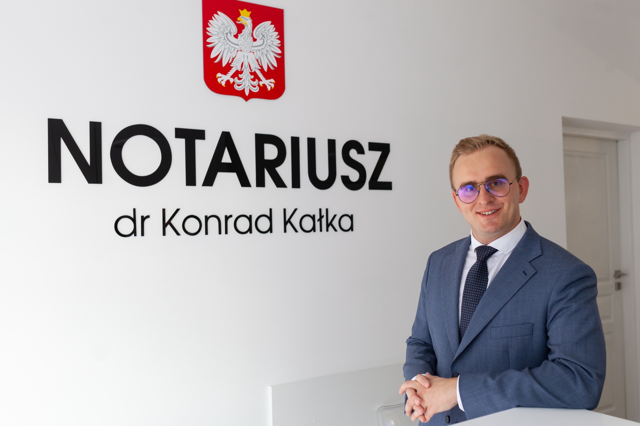 Uśmiechnięty mężczyzna w niebieskim garniturze i okularach, stojący na tle ściany z napisem 'NOTARIUSZ dr Konrad Katka' i godłem Polski.