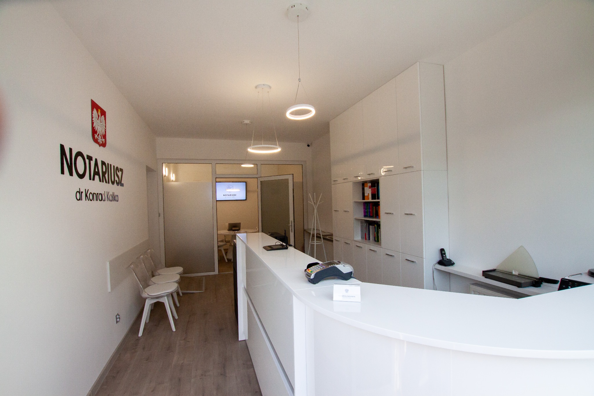 Jasne wnętrze kancelarii notarialnej z recepcją, logo z godłem Polski i nazwiskiem notariusza na ścianie, oraz widocznymi krzesłami dla klientów i oświetleniem w postaci okręgów.