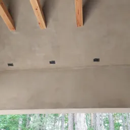 Poddasze z wykończoną gładzią, widoczne drewniane belki stropowe i kratki wentylacyjne, ściana z czerwonej cegły w prawym rogu, widok na drzewa na zewnątrz.