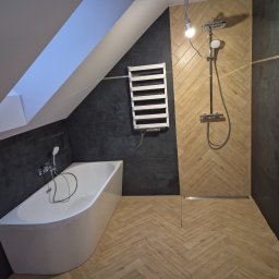Firma Remontowo Budowlana Mariusz Kondracki - Ukończona łazienka na poddaszu z wanną, grzejnikiem drabinkowym i prysznicem typu walk-in, wykończona drewnopodobnymi płytkami ułożonymi w jodełkę oraz ciemnymi płytkami na ścianach, z widoczną...