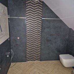 Firma Remontowo Budowlana Mariusz Kondracki - Ciemne płytki ścienne z dekoracyjnym pasem w jodełkę, jasna podłoga ułożona w jodełkę oraz biała toaleta w łazience ze skośnym sufitem.