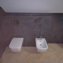 Firma Remontowo Budowlana Mariusz Kondracki - Nowoczesna łazienka z szarą, teksturowaną glazurą, białym kompaktem WC i bidetem, oraz podłogą w jasny parkiet ułożony w jodełkę.