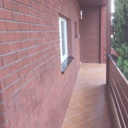 Firma Remontowo Budowlana Mariusz Kondracki - Wykończony balkon z czerwoną cegłą na ścianie, brązowymi płytkami na podłodze ułożonymi w karo, białym oknem z czarnym parapetem i nowoczesnym oświetleniem zewnętrznym.