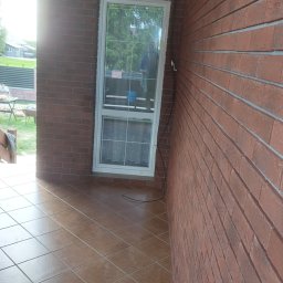 Firma Remontowo Budowlana Mariusz Kondracki - Zadaszony taras z brązowymi płytkami podłogowymi i ścianami z cegły klinkierowej, widoczne okno z białymi ramami i firankami. Na ścianie zewnętrzna lampa.