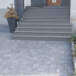 Firma Remontowo Budowlana Mariusz Kondracki - Wejście do domu z szarymi schodami wyłożonymi płytkami, brązowe drzwi z pionowym przeszkleniem, elewacja z szarego kamienia, obok schodów donica z uschniętą choinką, podjazd wyłożony szarą kostką.