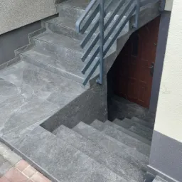 Zewnętrzne schody wyłożone szarą płytką imitującą kamień, z metalową balustradą, prowadzące do brązowych drzwi wejściowych.