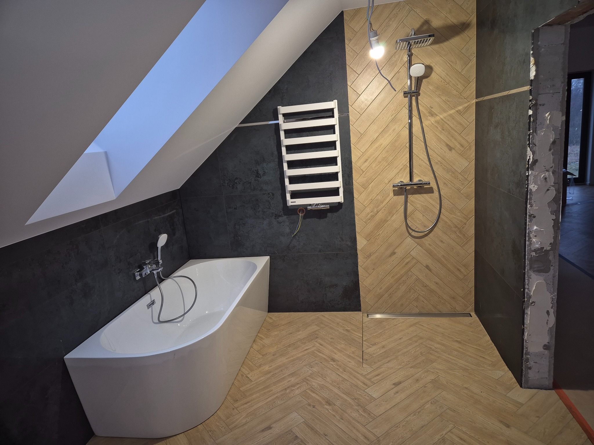 Ukończona łazienka na poddaszu z wanną, grzejnikiem drabinkowym i prysznicem typu walk-in, wykończona drewnopodobnymi płytkami ułożonymi w jodełkę oraz ciemnymi płytkami na ścianach, z widoczną...