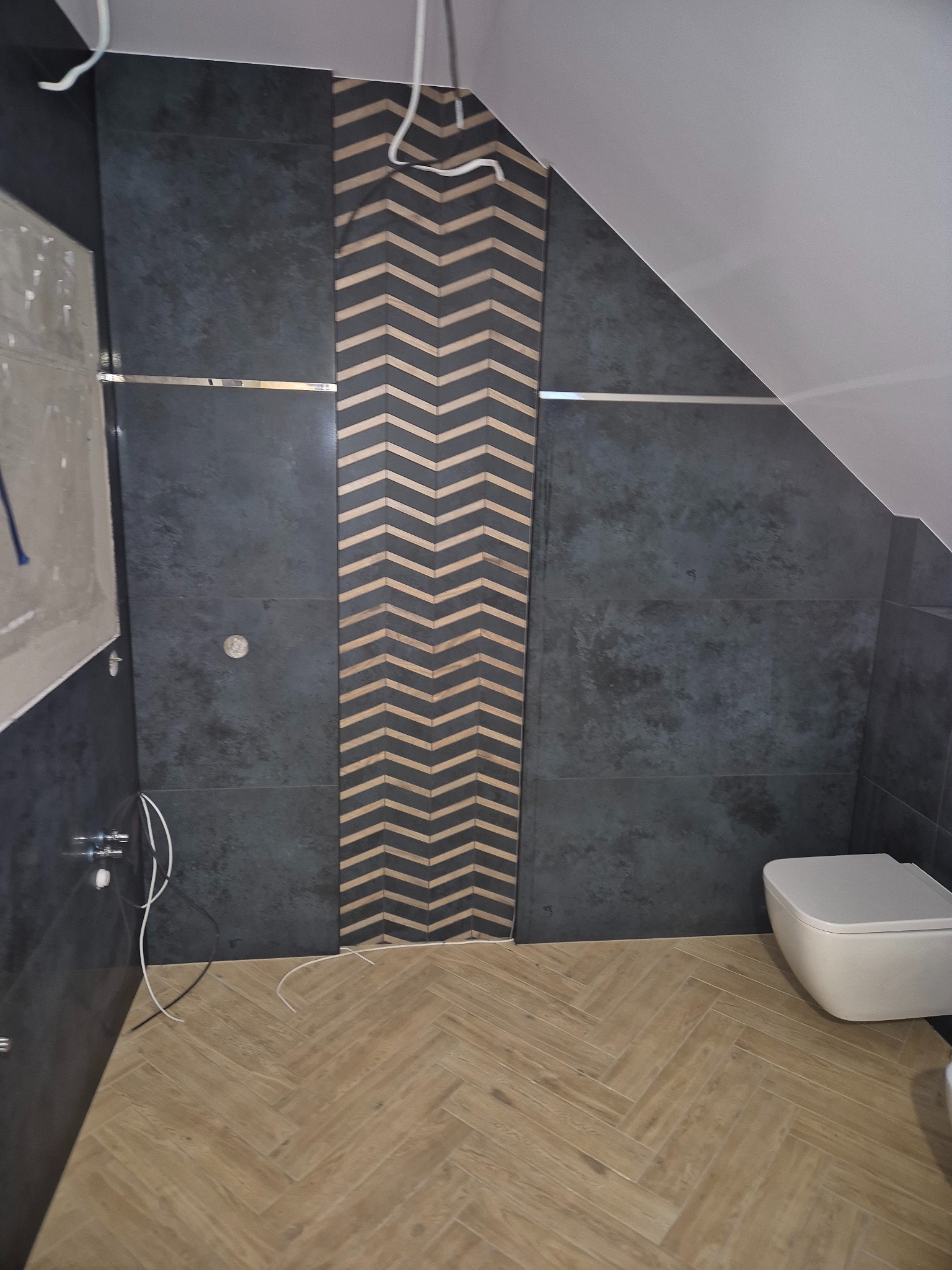 Ciemne płytki ścienne z dekoracyjnym pasem w jodełkę, jasna podłoga ułożona w jodełkę oraz biała toaleta w łazience ze skośnym sufitem.