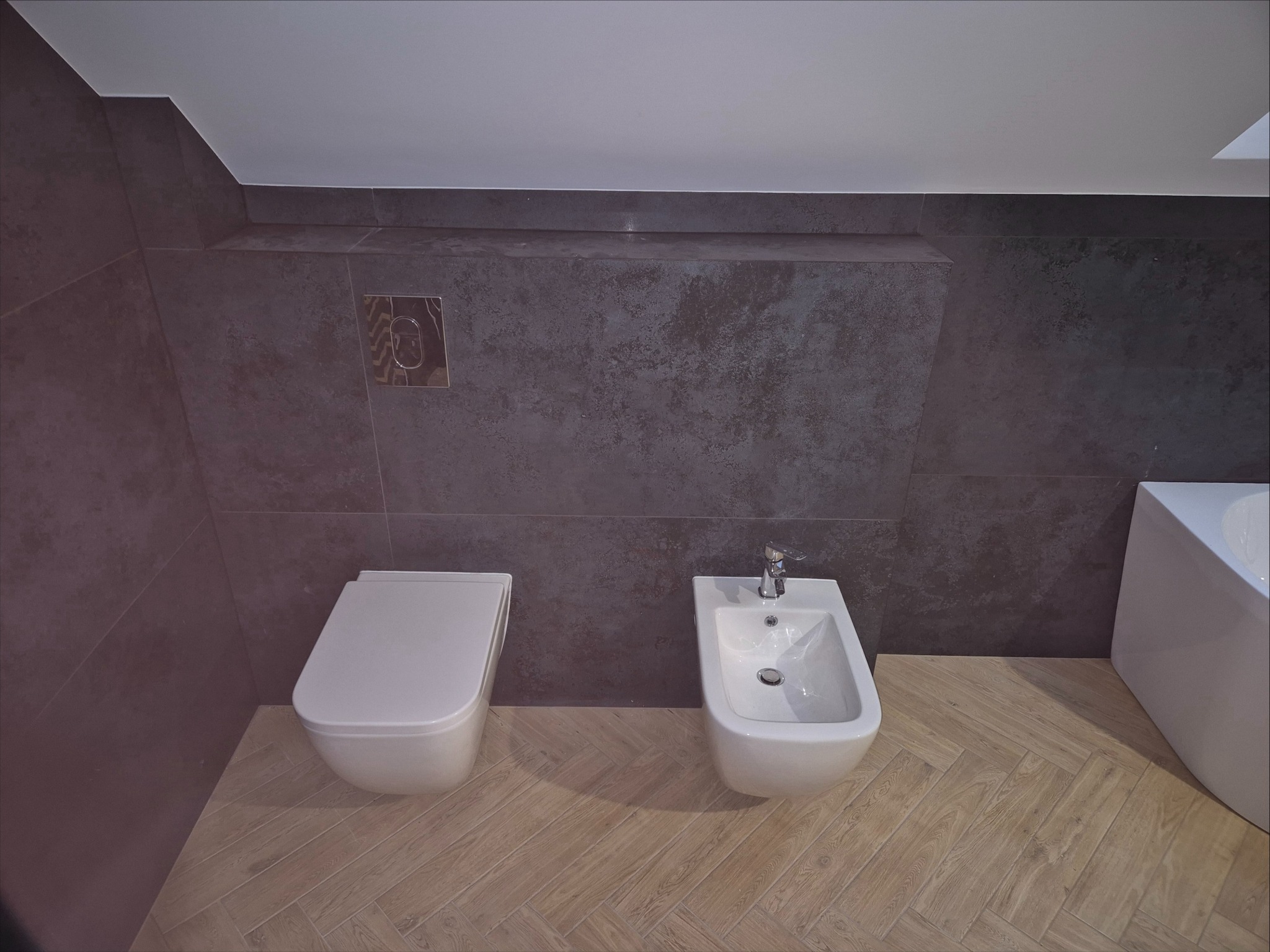 Nowoczesna łazienka z szarą, teksturowaną glazurą, białym kompaktem WC i bidetem, oraz podłogą w jasny parkiet ułożony w jodełkę.