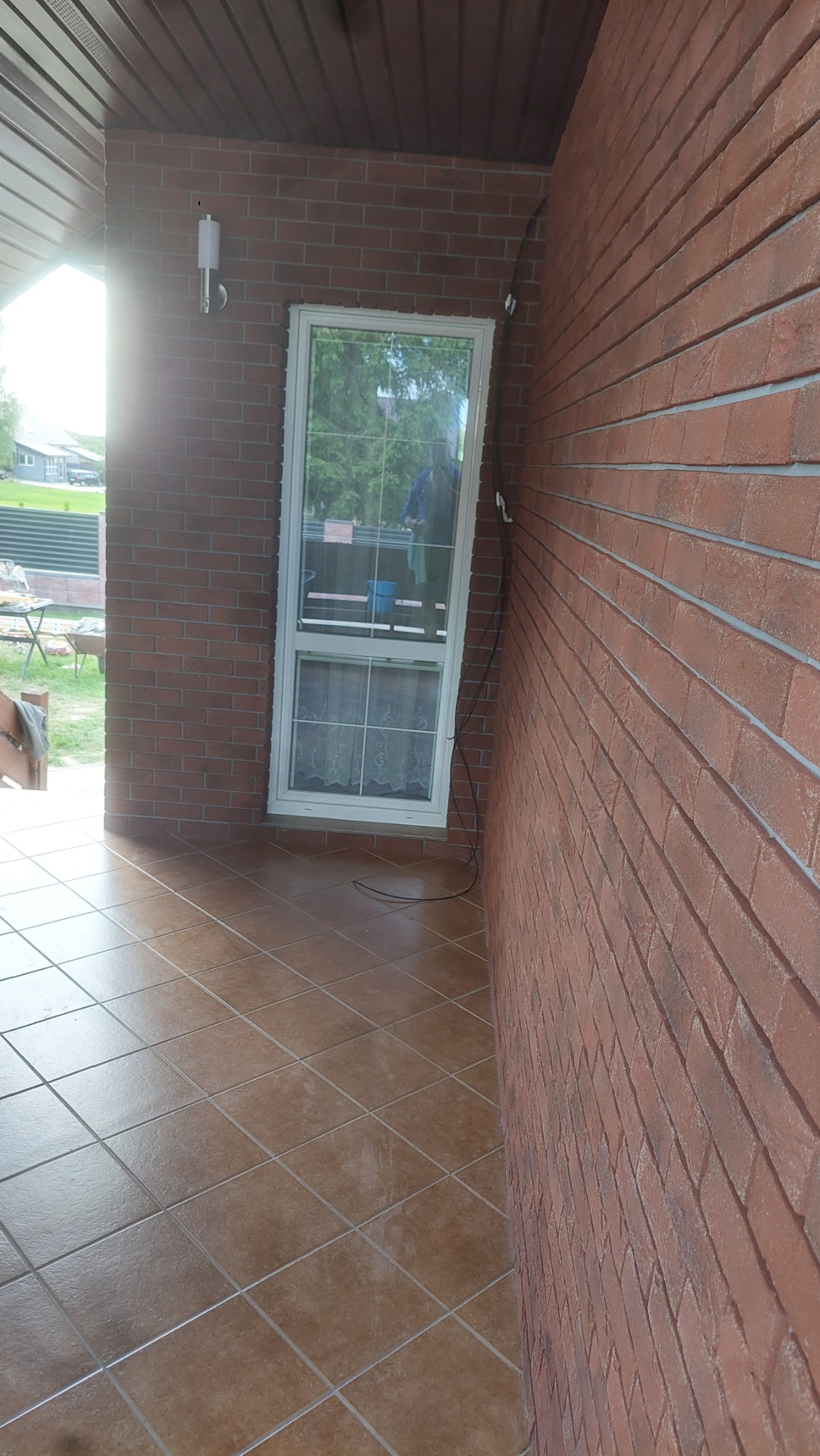 Zadaszony taras z brązowymi płytkami podłogowymi i ścianami z cegły klinkierowej, widoczne okno z białymi ramami i firankami. Na ścianie zewnętrzna lampa.