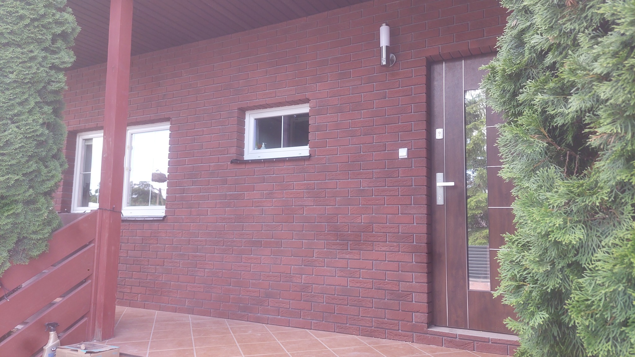 Ceglana elewacja budynku z brązowymi drzwiami wejściowymi z elementami szkła, dwoma oknami z białymi ramami i zewnętrzną lampą ścienną, częściowo zasłonięta przez zielone krzewy.