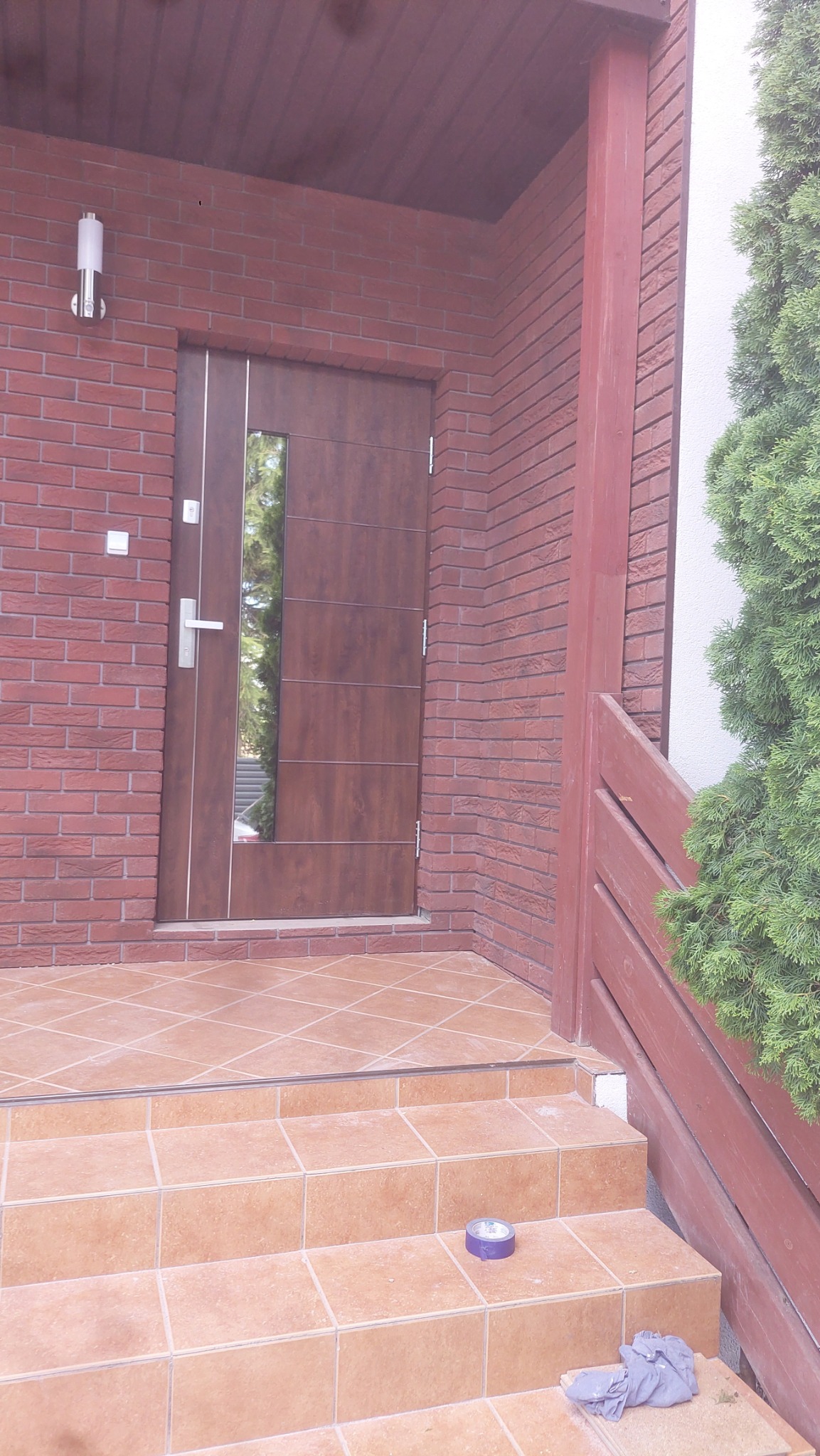 Wejście do domu z brązowymi drzwiami z szybą, wykończone czerwoną cegłą, z ceramicznymi schodami i podłogą, na których leżą niebieska taśma i szara szmatka.