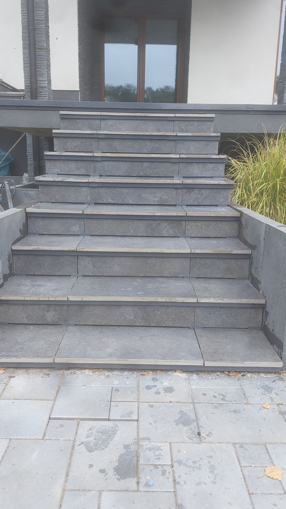 Schody zewnętrzne wyłożone szarą płytką, z widocznymi elementami montażowymi, prowadzące do domu z oknem odbijającym drzewa.