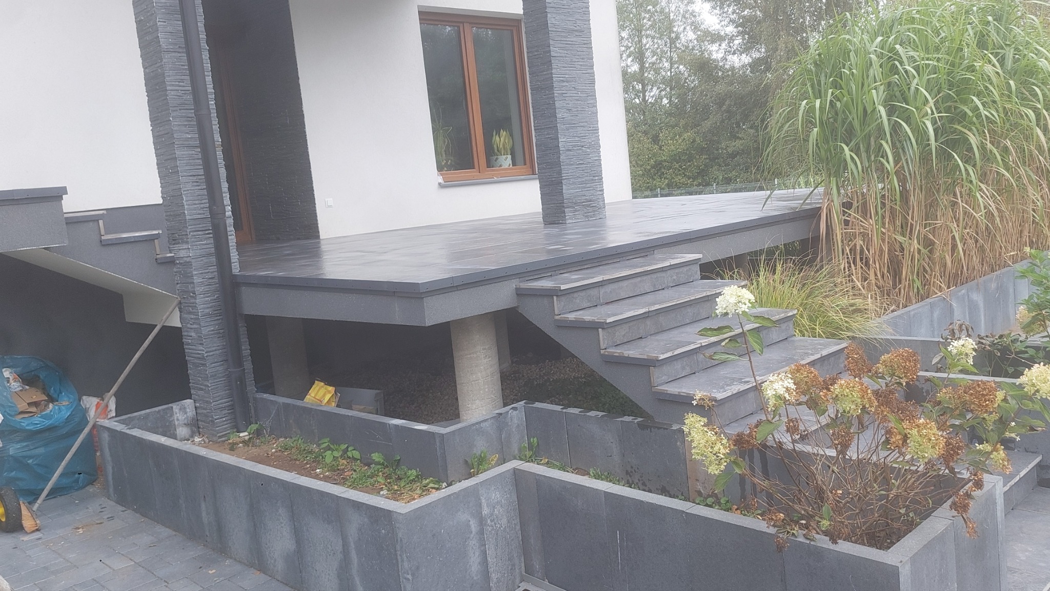 Szara, nowoczesna elewacja domu z tarasem, schodami i betonowymi donicami z roślinami, widok z góry na fragment elewacji.