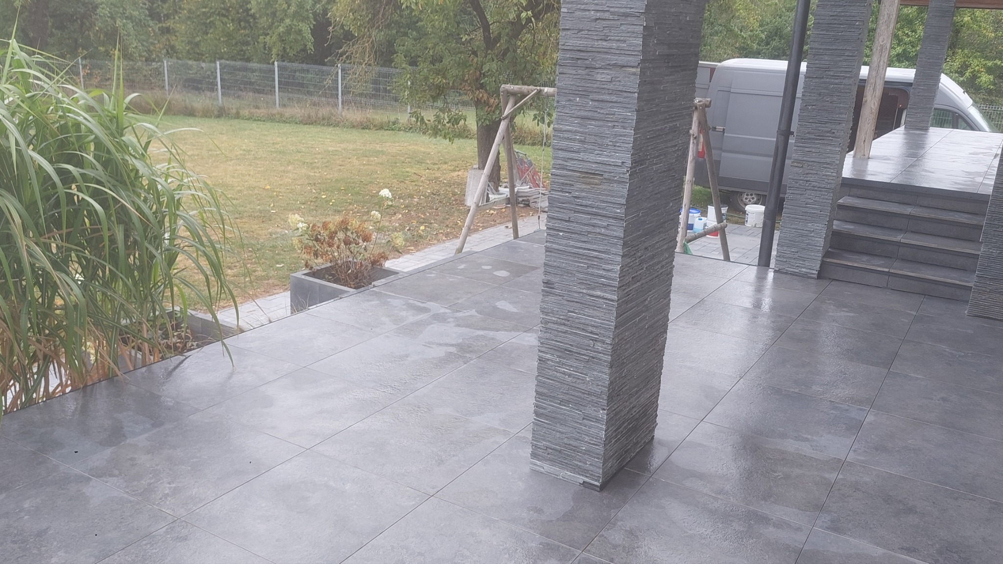 Wyłożony szarą płytką taras z kolumnami obłożonymi kamieniem łupkowym, widok na ogród z huśtawką i zaparkowany samochód dostawczy w tle.