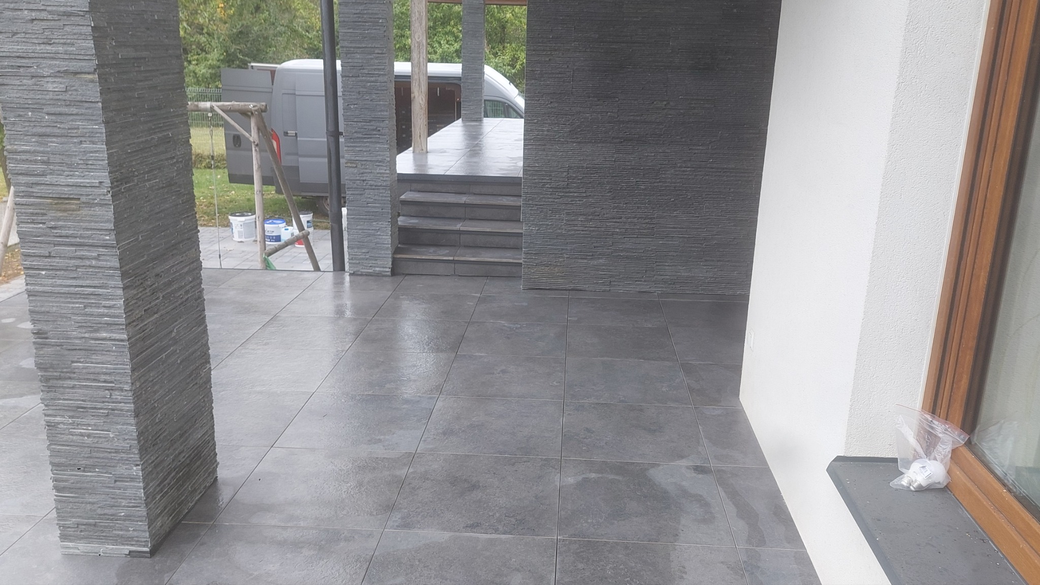 Szare płytki podłogowe na tarasie z kolumnami obłożonymi kamieniem łupkowym, widoczne schody i fragment białej ściany z oknem, na parapecie leży zapakowana żarówka LED.